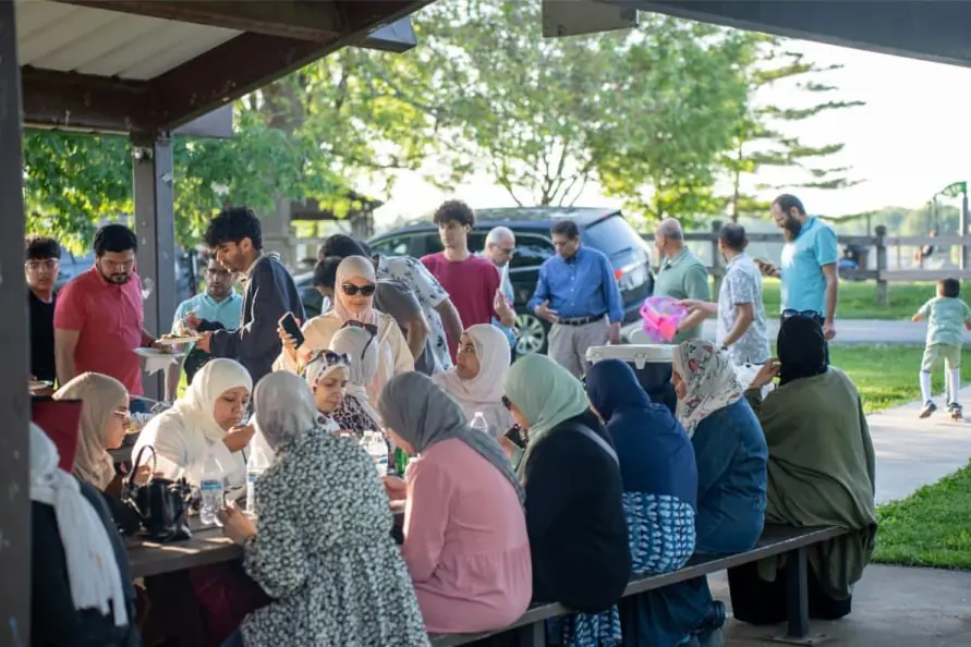 Families sharing a meal at long tables under a park shelter at the MAS Family Potluck